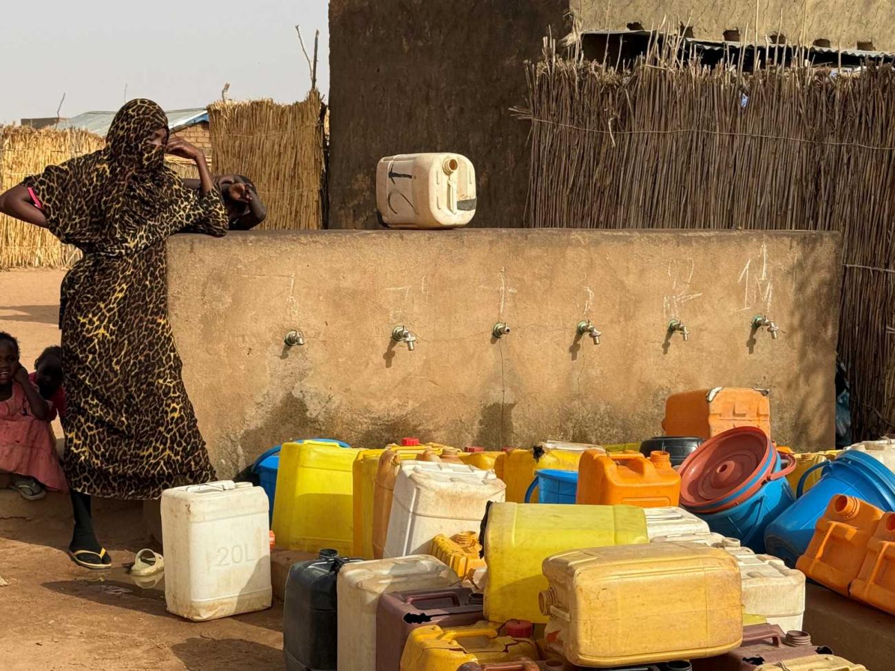 A woman in patterned clothing stands by a water point with multiple taps, with many empty jerrycans and buckets in front.