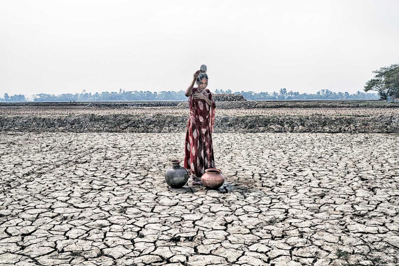 A woman in a red sari stands on cracked, dried ground and pours water over her head; two large water pots sit beside her.
