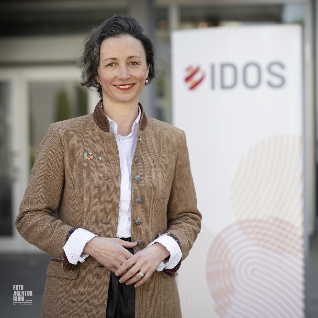 Portrait of a smiling woman in a brown blazer standing before a blurred “IDOS” banner, hands loosely clasped in front.