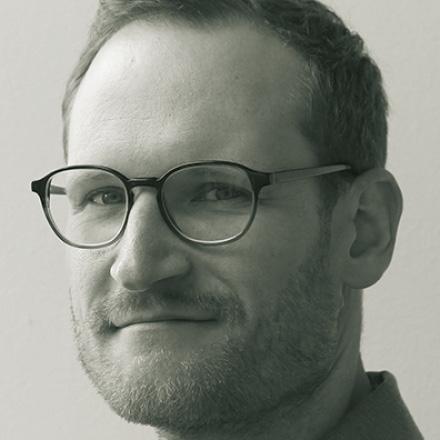 mit Brille und Bartstoppeln, der leicht lächelt und seitlich in die Kamera blickt.	Black-and-white portrait of a man with glasses and light stubble, slightly smiling and looking toward the camera from the side.