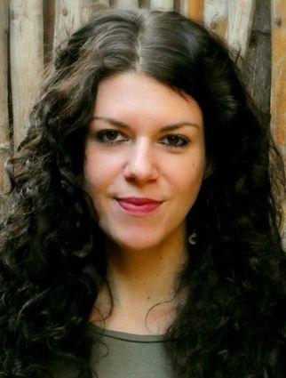 Portrait of a woman with long, dark curly hair in front of a weathered wooden wall, looking directly at the camera.