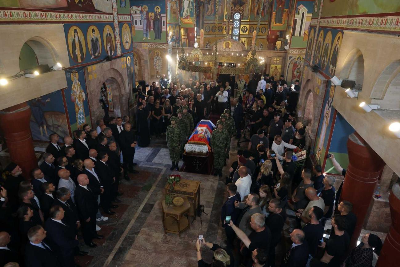 A coffin draped with a flag stands inside an Orthodox church; six soldiers stand around the coffin, and a crowd of people is gathered in the church.