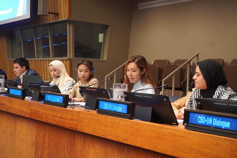 Several young people are seated in a row on a panel; in front of them are microphones and signs bearing the words “SCO-UN Dialogue”.