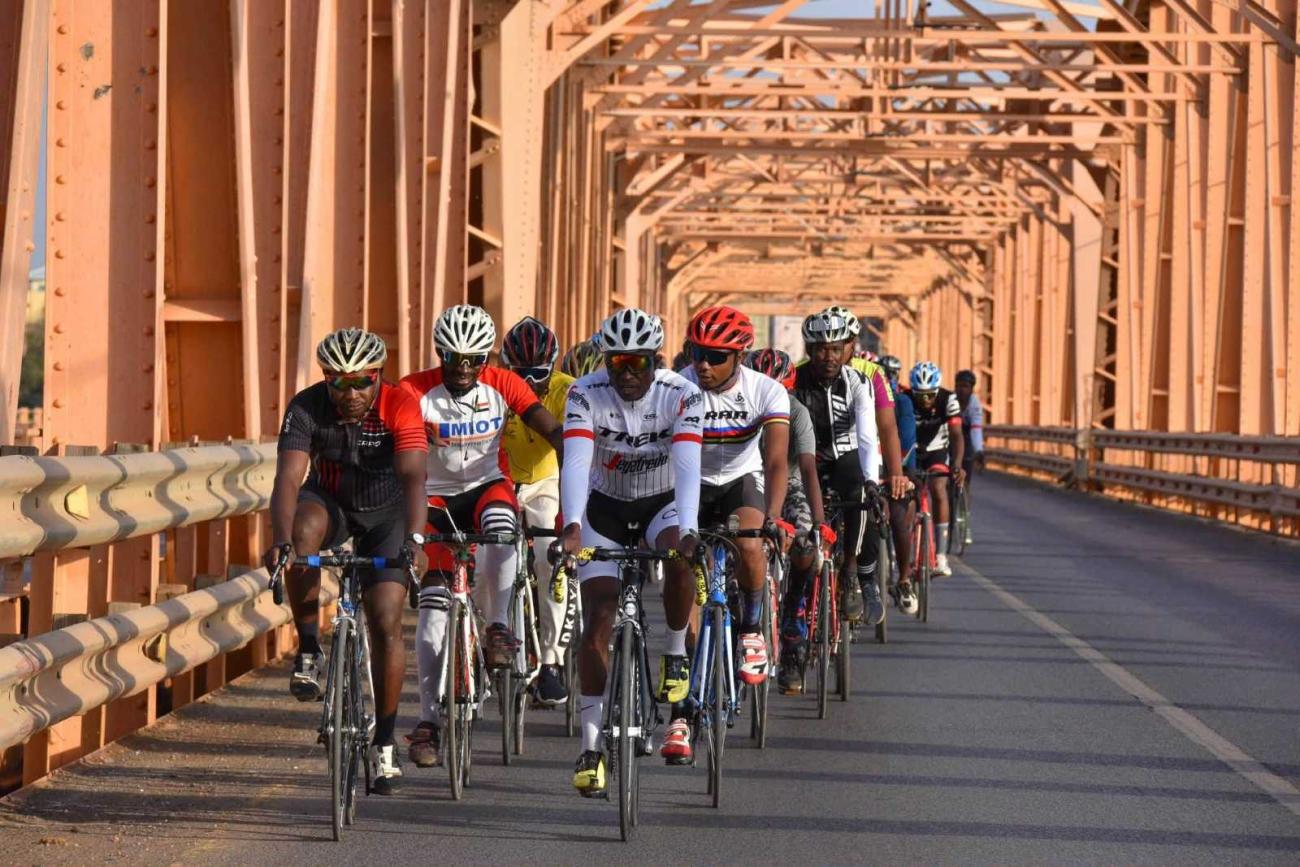 A group of helmeted road cyclists rides in a line across a steel bridge in warm evening light.