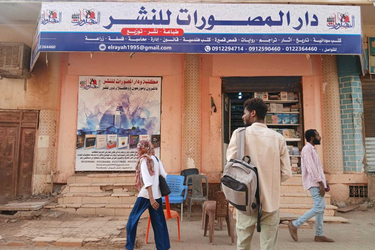 Storefront of the Dar Al-Massoorat publishing house and bookshop in Sudan, with Arabic signage, bookshelves and passing pedestrians.