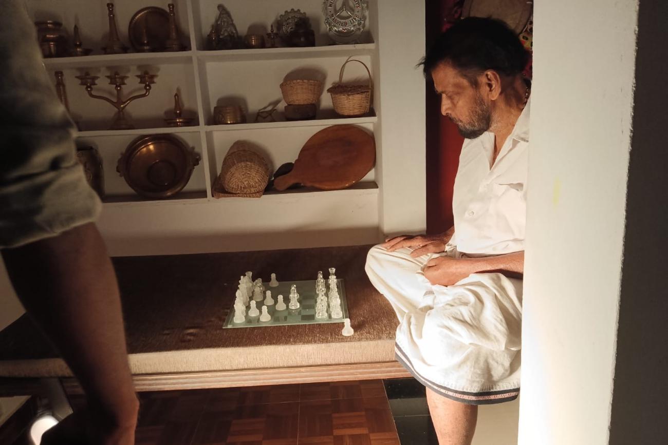 A man in white sits side-on on a bench, studying a chessboard with glass pieces; shelves of vessels and baskets are in the background. 