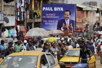 picture alliance/AP Photo/Sunday Alamba Belebte Straßenszene in einem afrikanischen Markt mit zahlreichen Menschen, Verkaufsständen und einem großen Wahlplakat von Paul Biya im Hintergrund.